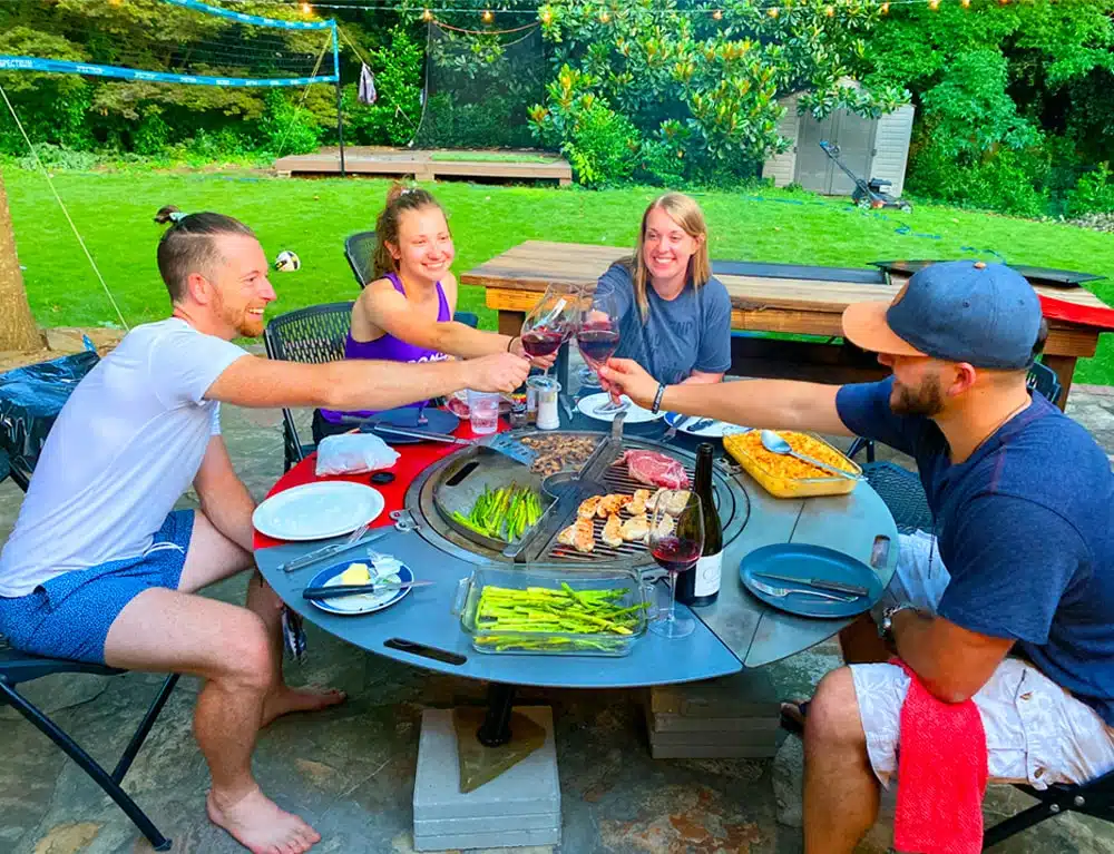 four people seated around a grill combo clinking their wine glasses together in a toast.
