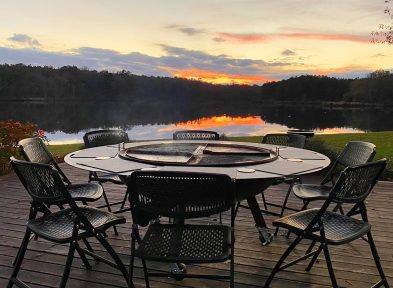 A gather grill combo surrounded by six chairs, on a patio by the lake, at sunset